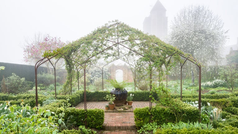 Misty foliage and new white flowers grow around a central arbour in a structured garden